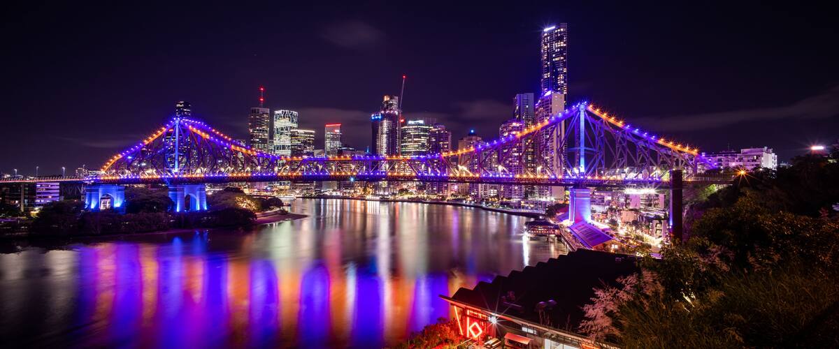 A brightly lit bridge with colorful lights reflecting on the water, surrounded by a city skyline with illuminated buildings.