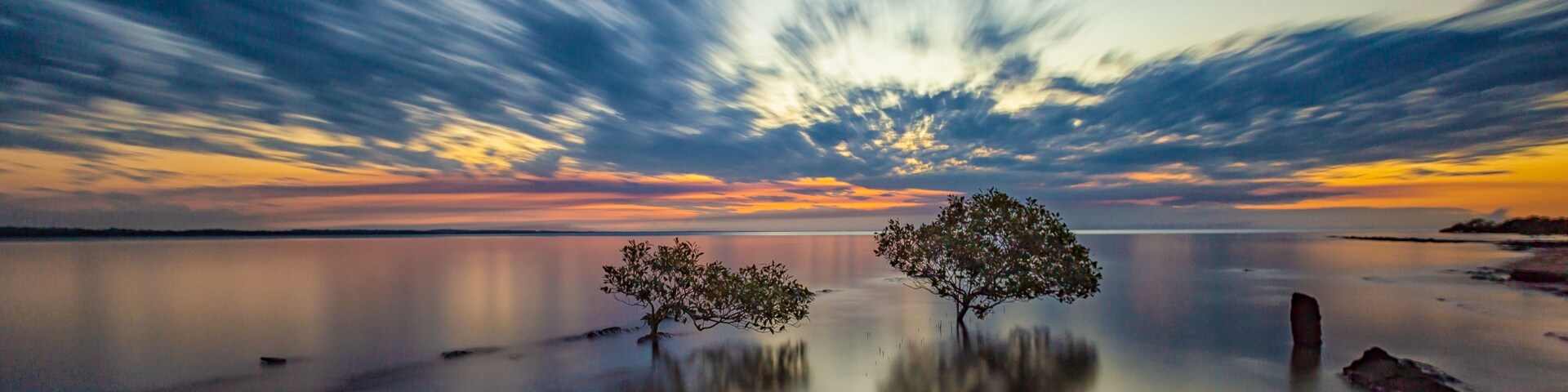 Um lago tranquilo com duas árvores em primeiro plano e um céu dramático com nuvens.