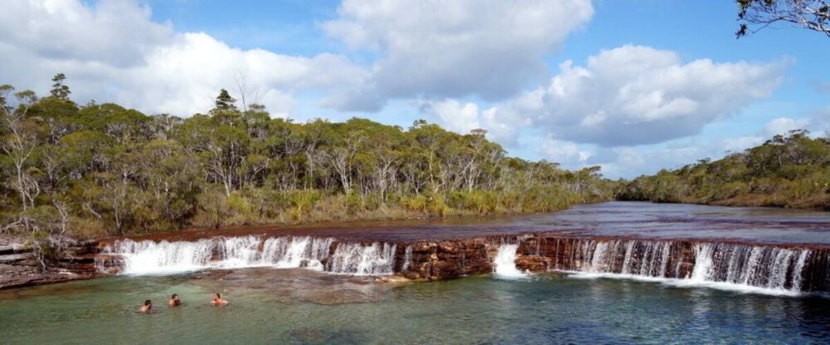 A clear water body with a waterfall, surrounded by trees and a cloudy sky.