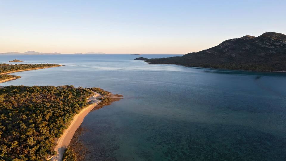 A coastal landscape with a sandy beach, clear blue waters, and a forested area.