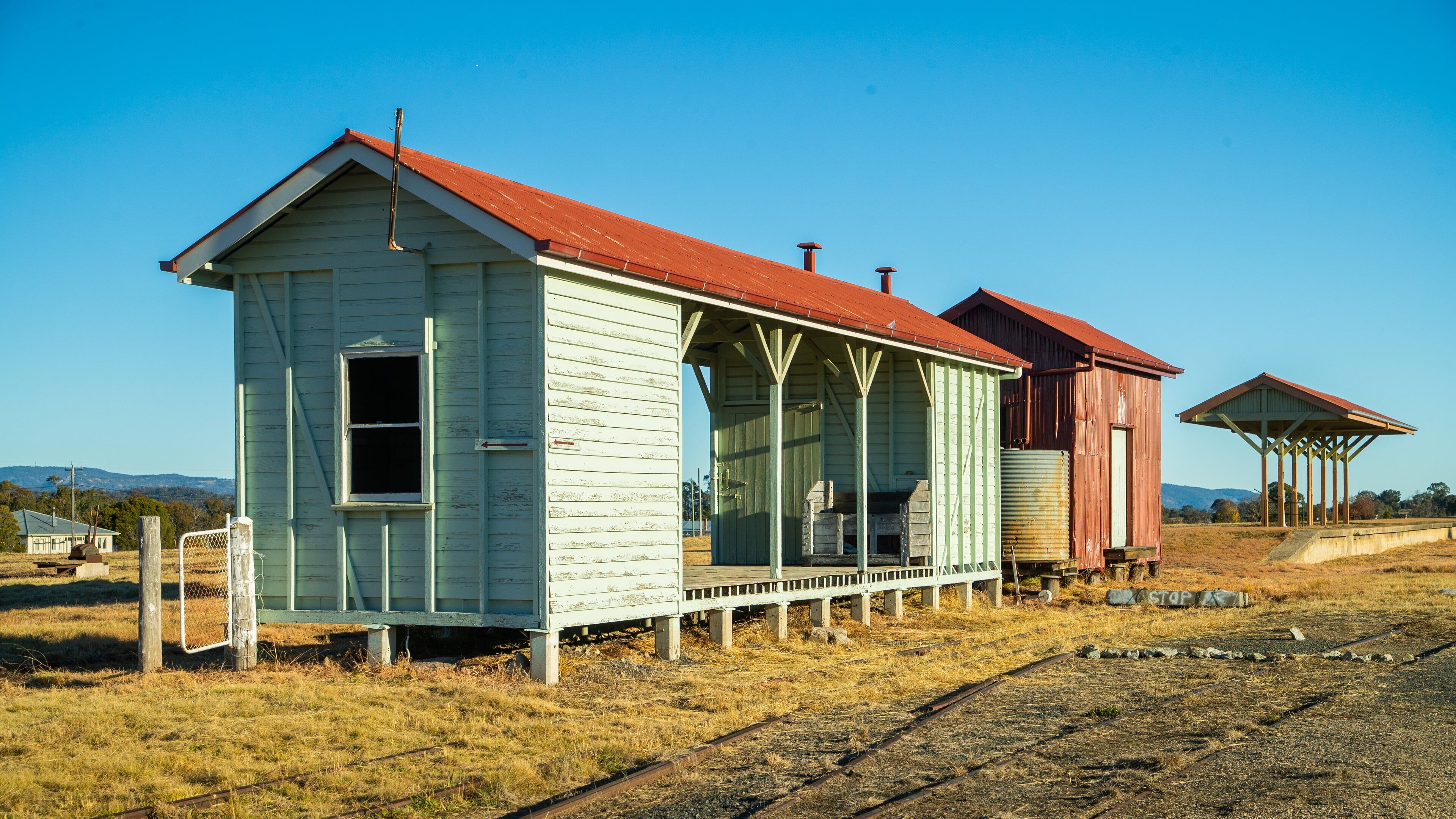 A small, weathered building with a red roof and a porch, surrounded by dry grass and a railway track.