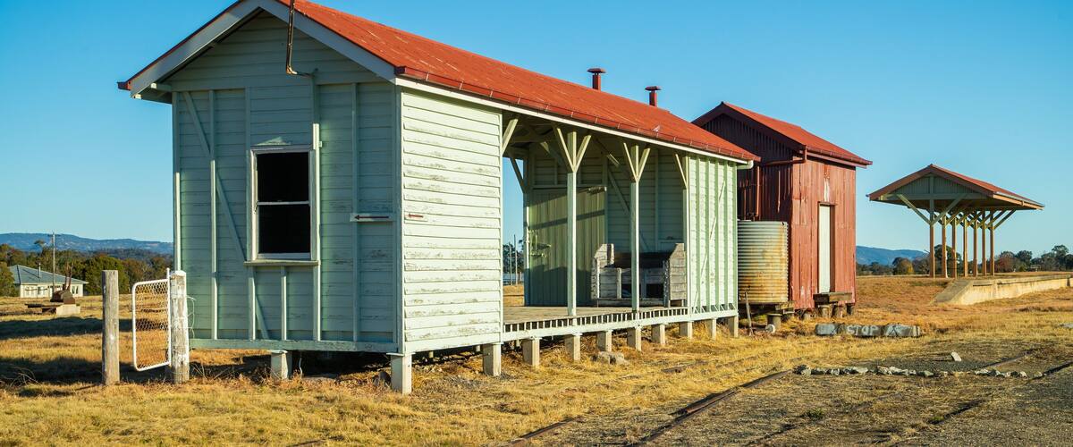 A small, weathered building with a red roof and a porch, surrounded by dry grass and a railway track.