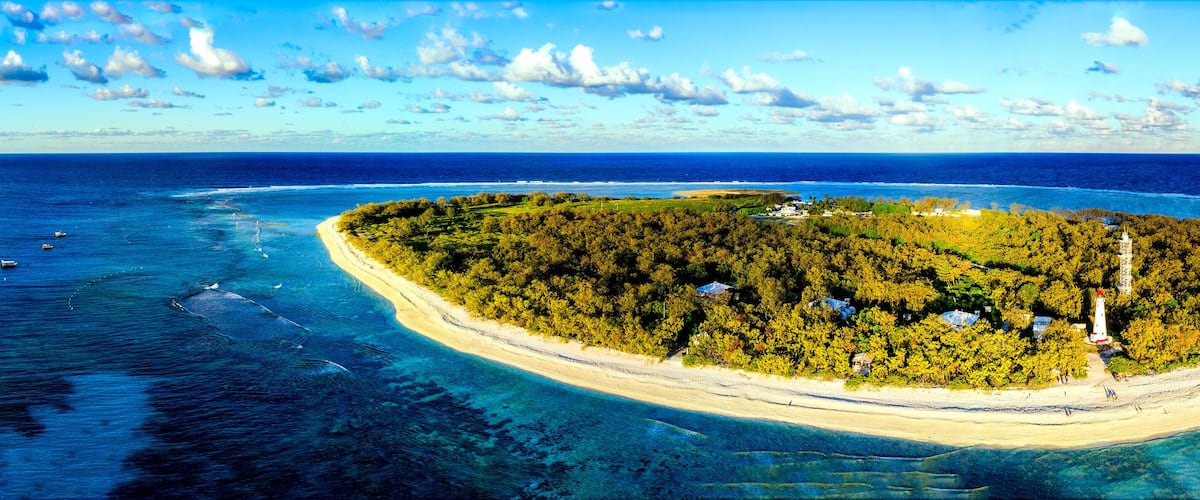 An island with a lighthouse, surrounded by clear blue waters and a sandy beach.