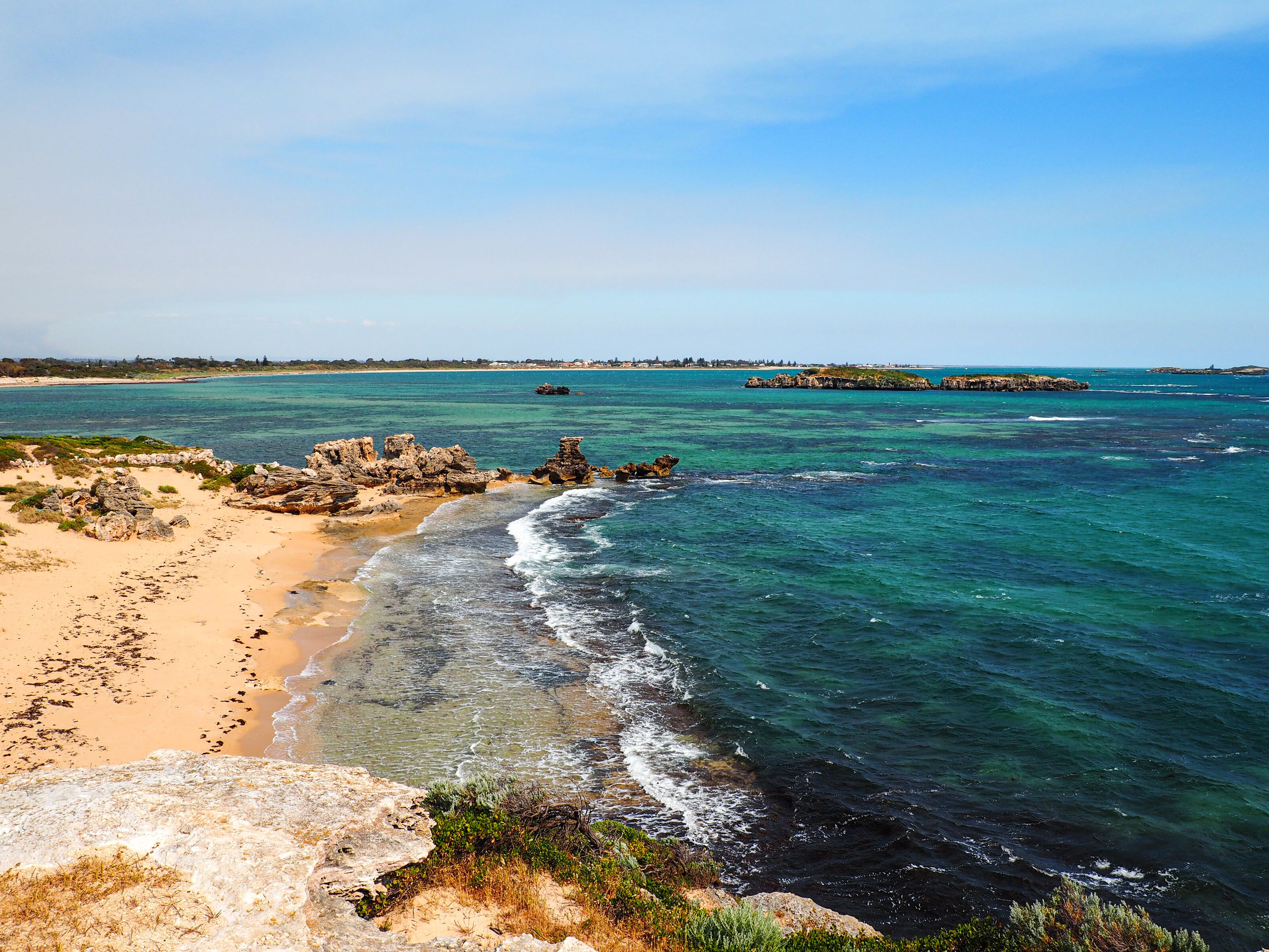 Paisagem costeira com águas turquesa cristalinas, praia de areia, afloramentos rochosos e formações terrestres ao longe.