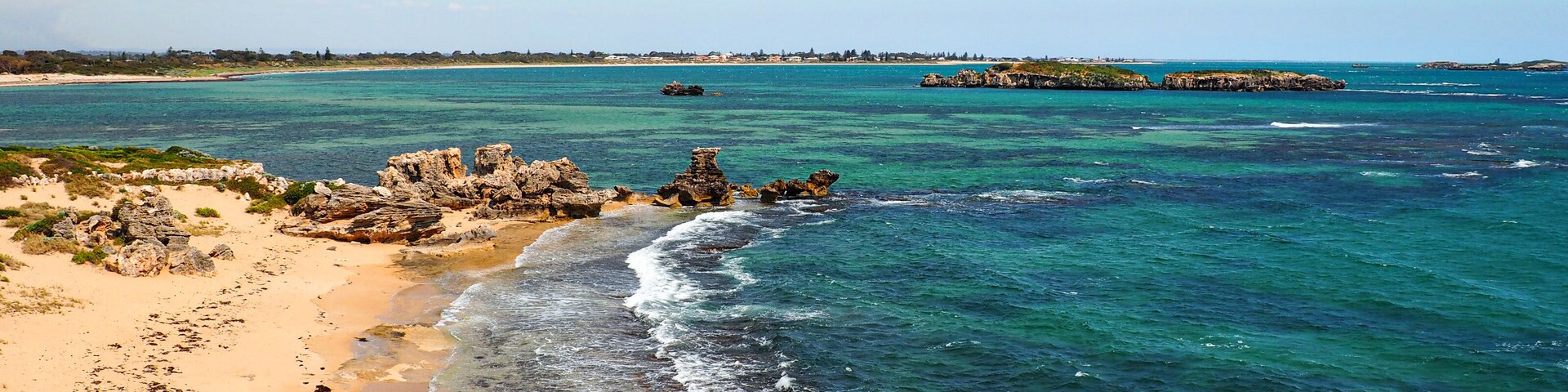 Paisagem costeira com águas turquesa cristalinas, praia de areia, afloramentos rochosos e formações terrestres ao longe.
