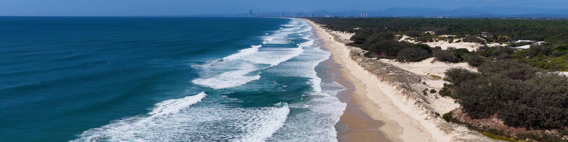 A coastal landscape with a sandy beach, ocean waves, and a distant city skyline.