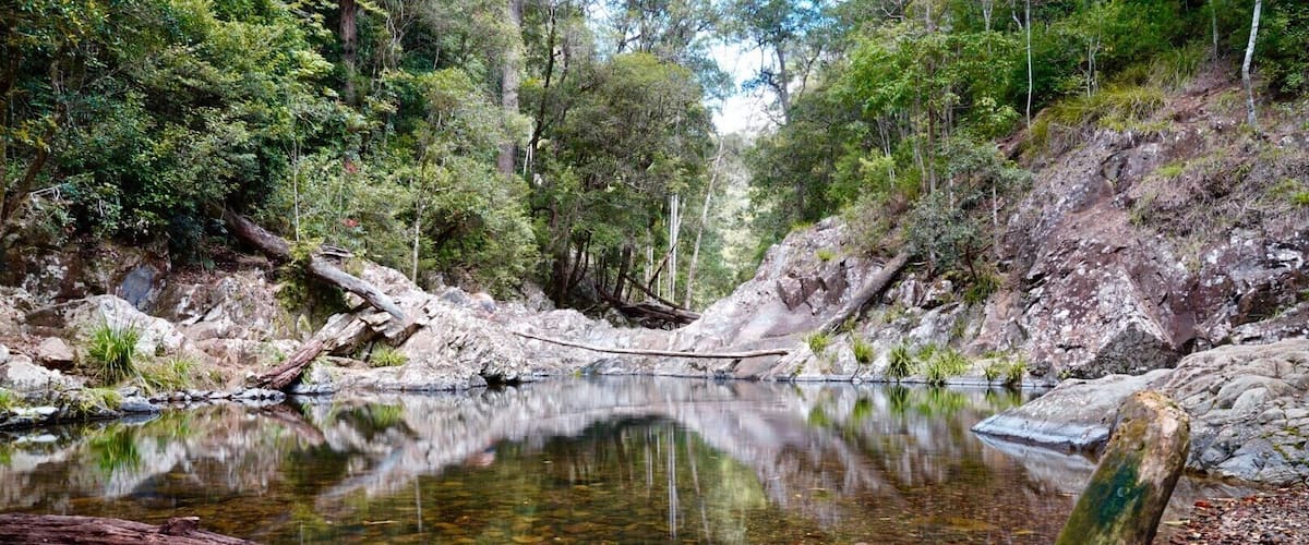 A calm river with clear water, surrounded by dense forest and rocky terrain.