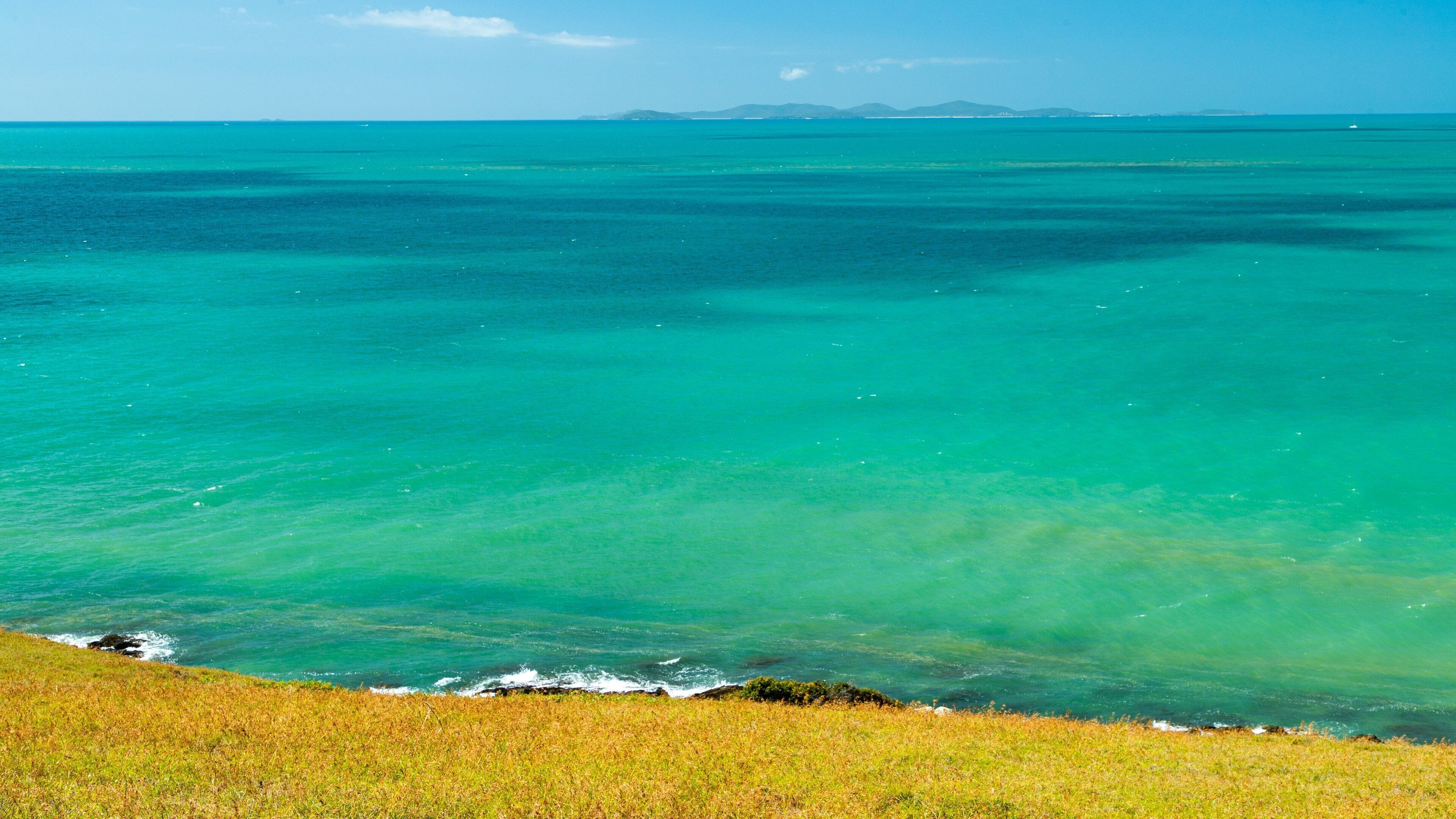 A coastal landscape with clear turquoise waters, a grassy foreground, and distant mountains under a clear sky.