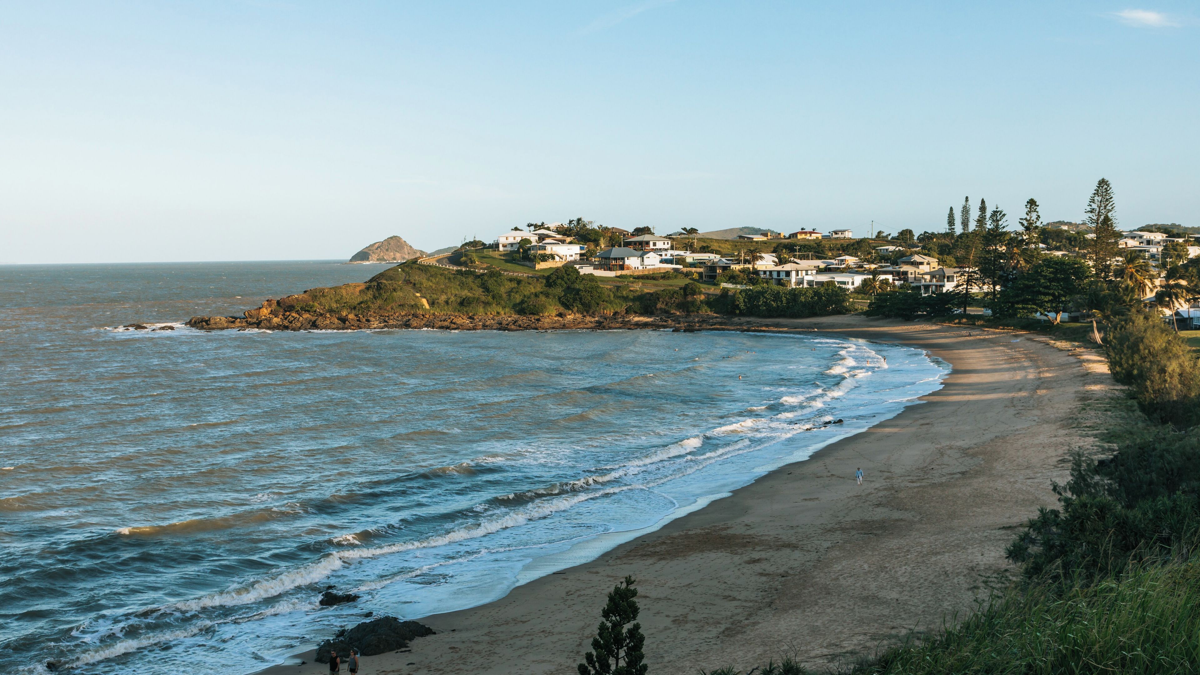 A coastal landscape with a sandy beach, ocean waves, and a residential area with houses on a hillside.