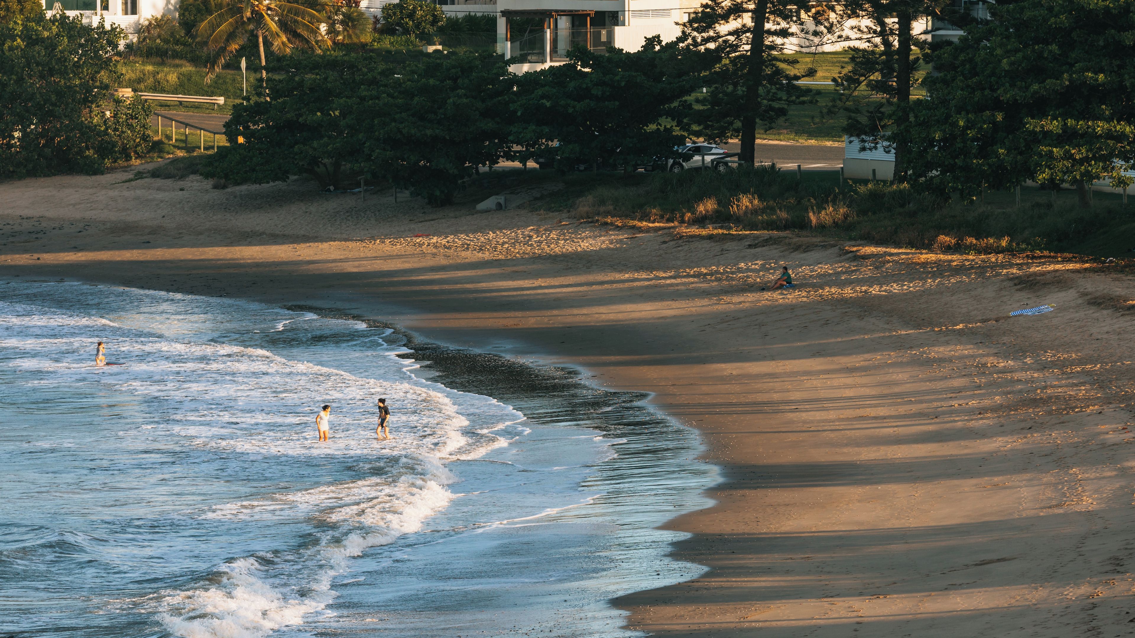 A beach with waves, a person sitting on the sand, and a building in the background.