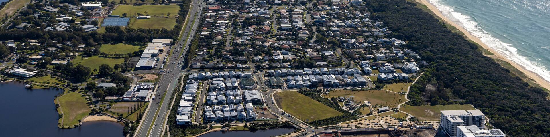 An aerial view of a coastal city with a long beachfront, dense residential areas, and a river running parallel to the coast.