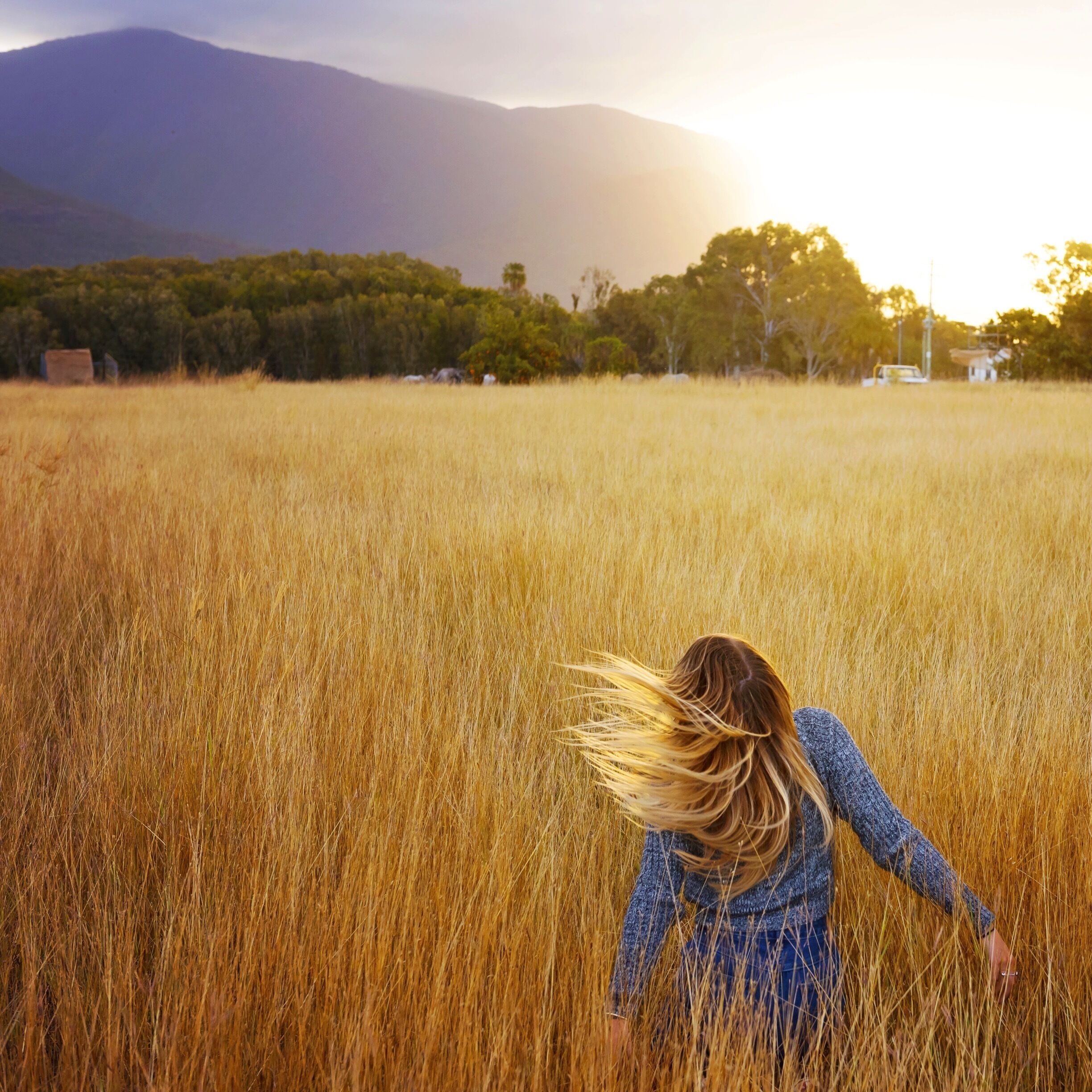 A woman in a field with tall golden grass, mountains in the background, and trees.