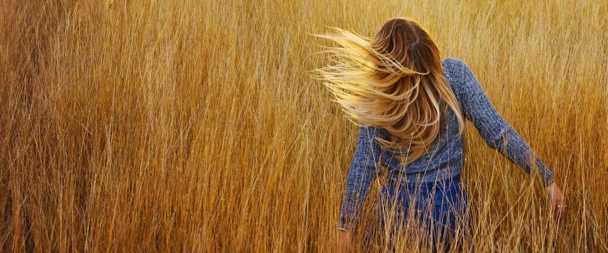 A woman in a field with tall golden grass, mountains in the background, and trees.