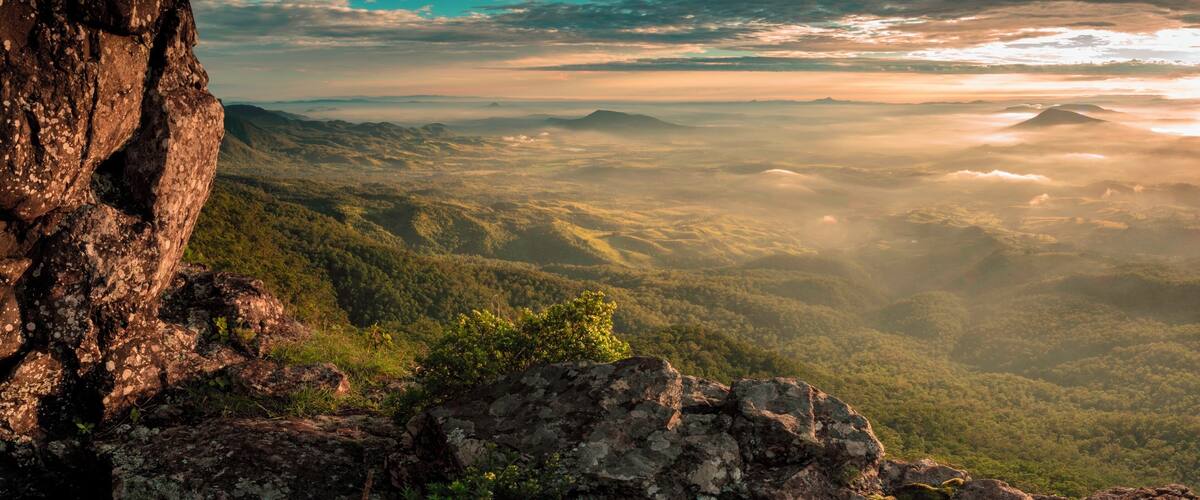 A mountainous landscape with a prominent rocky outcrop in the foreground and a vast forested valley beneath.