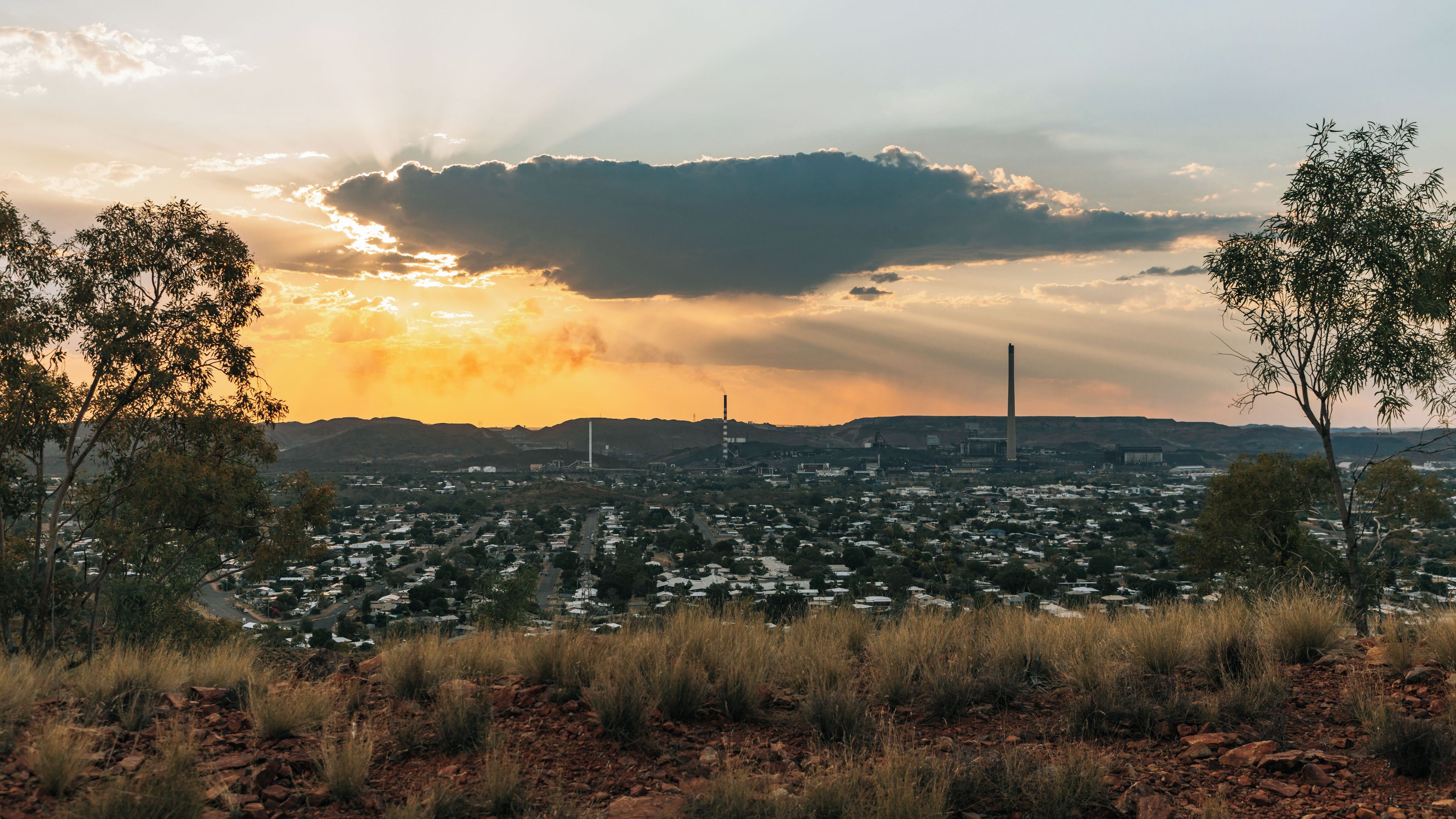 A cityscape at sunset with a prominent factory chimney, a dense residential area, and a foreground of dry grass and red rocks.