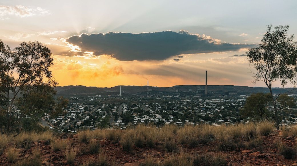A cityscape at sunset with a prominent factory chimney, a dense residential area, and a foreground of dry grass and red rocks.