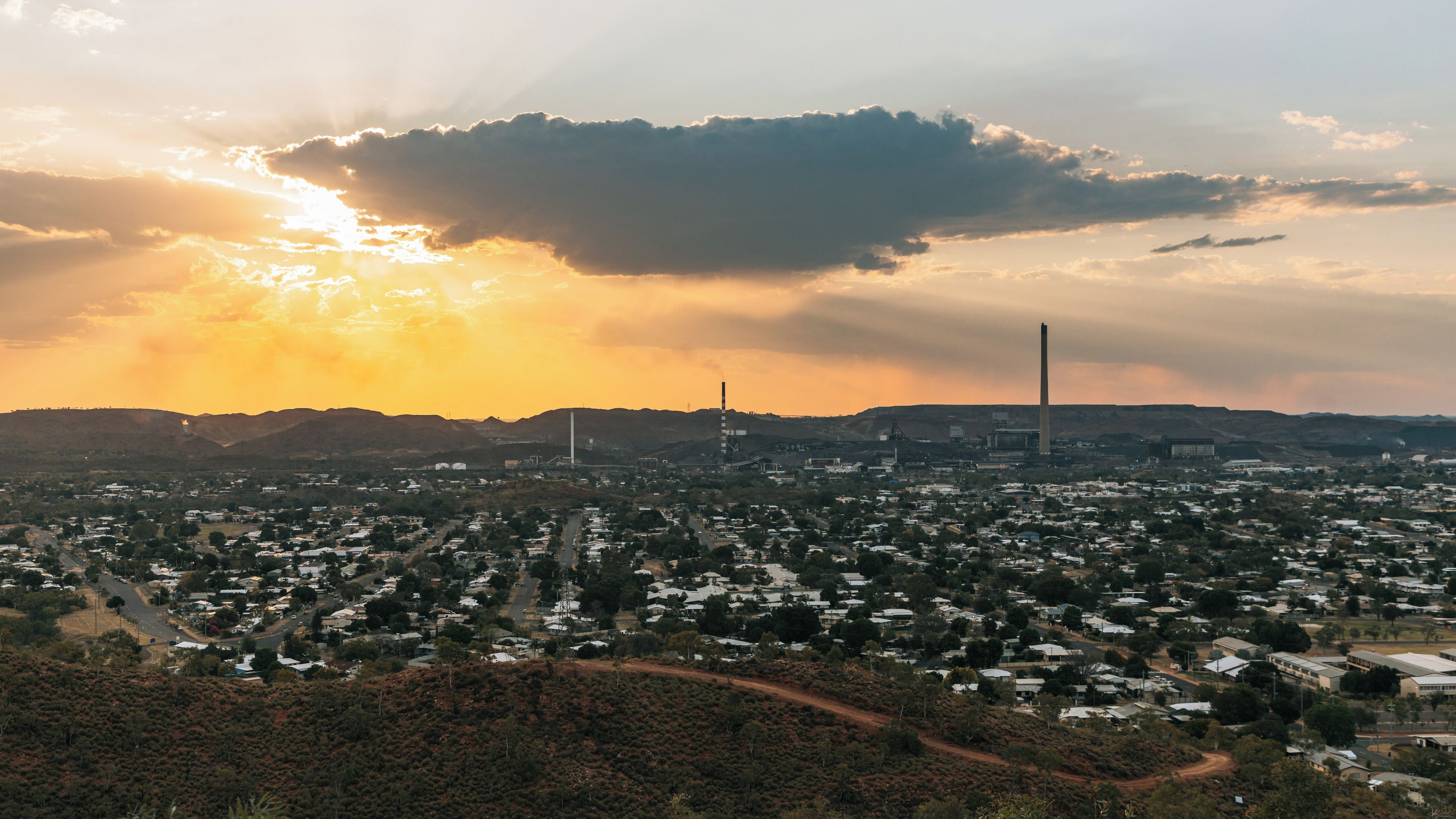 Un paysage urbain avec une zone industrielle bien visible, comprenant de hautes cheminées et une grande usine, le tout baigné par la lumière spectaculaire d’un coucher de soleil.