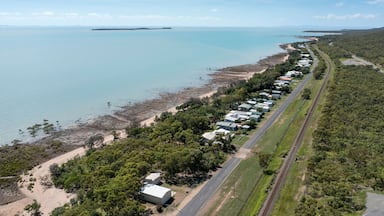 An aerial view of a coastal area with a row of mobile homes along a road, a beach, and a clear blue sea.