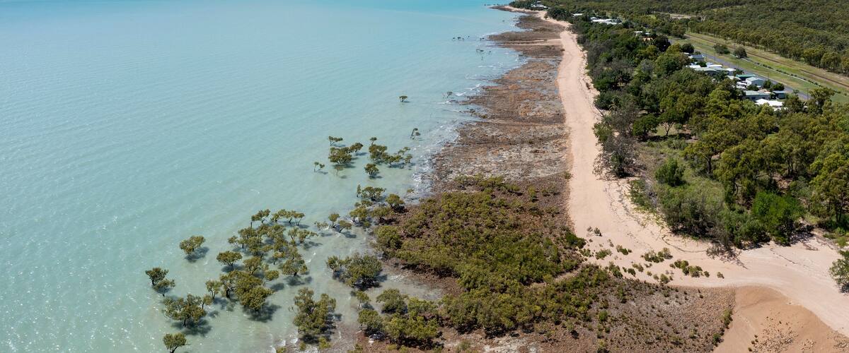 A coastal landscape with a sandy beach, clear turquoise waters, and dense green vegetation.