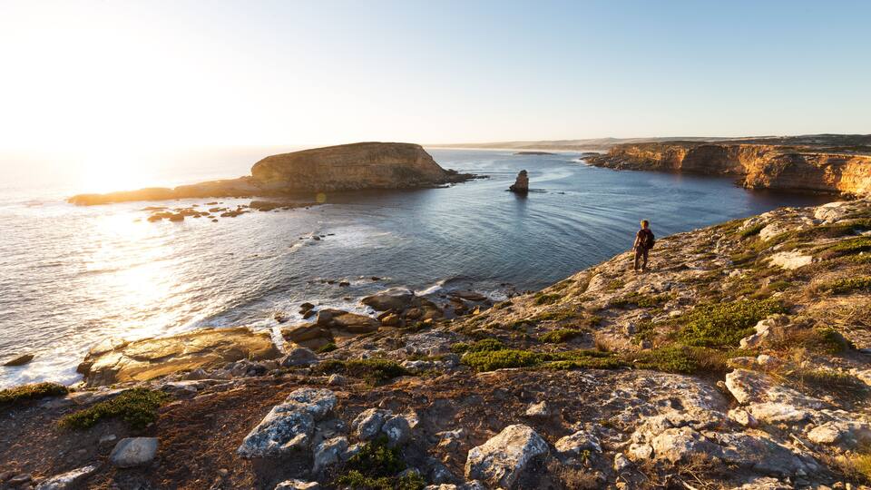 A rocky coastline with a person standing on a cliff overlooking the sea, a prominent rock formation in the distance, and a clear sky.