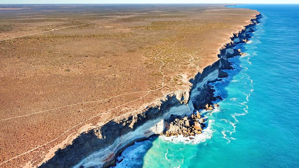 A coastal landscape with a rocky cliff edge, a clear blue ocean, and a flat, arid terrain.