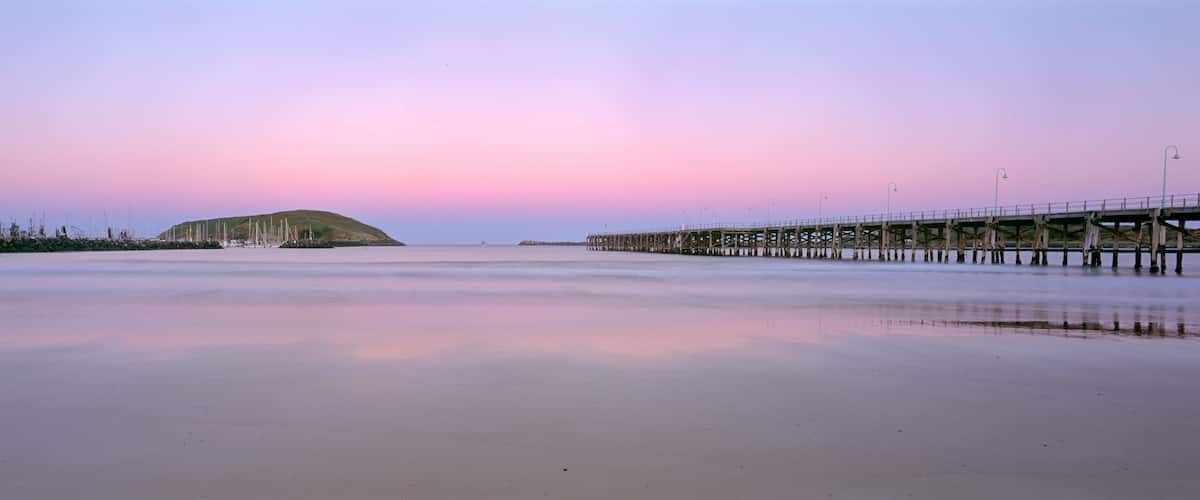 A beach with a pier extending into the water, a hill in the distance, and a sunset sky.
