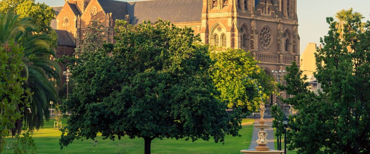 A cathedral with two prominent spires, surrounded by lush green trees and a well-maintained garden with a fountain.