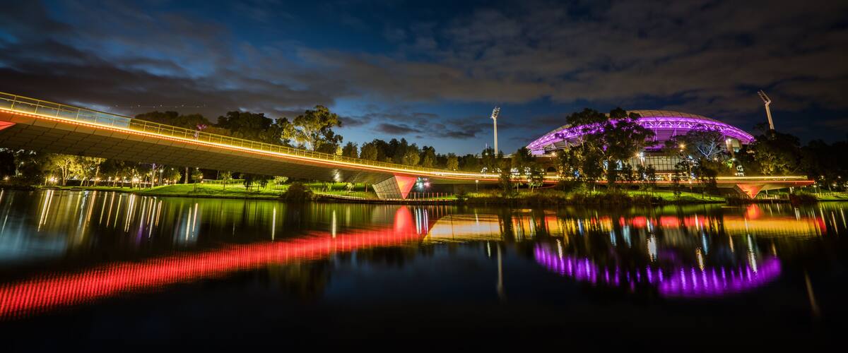 A nighttime view of a bridge with lights reflecting on a body of water, a stadium with purple lighting, and a tall structure with lights.