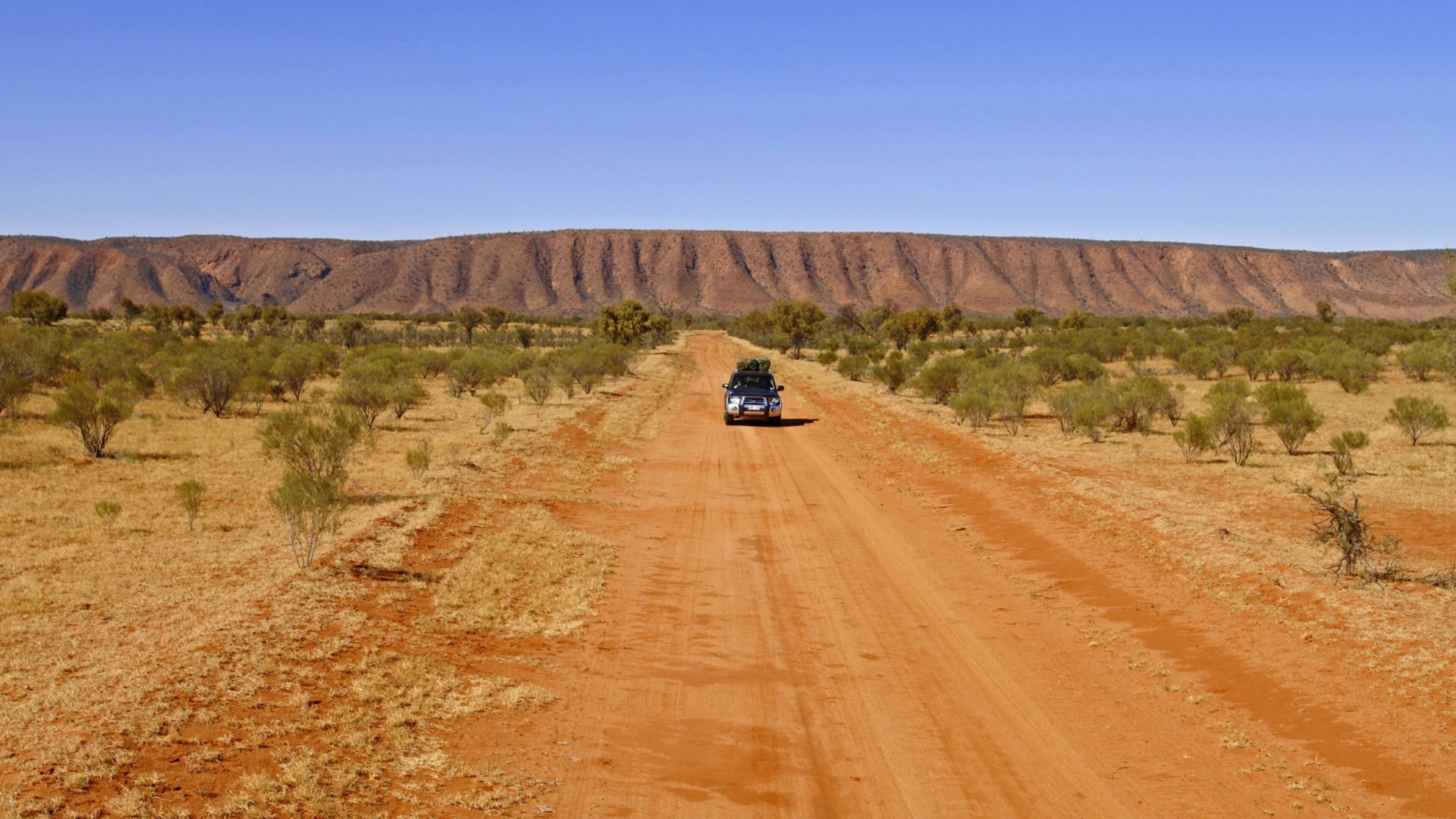 A vehicle on a dirt road in a desert landscape with sparse vegetation and a mountain range in the background.