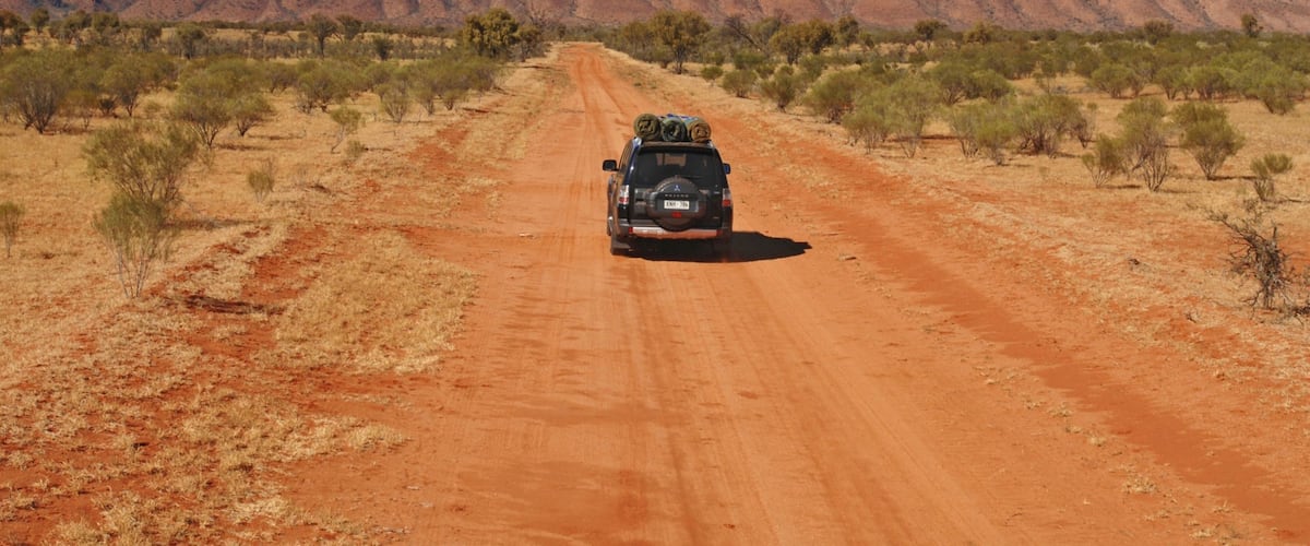 A red dirt road with sparse vegetation and a large red rock formation in the distance.