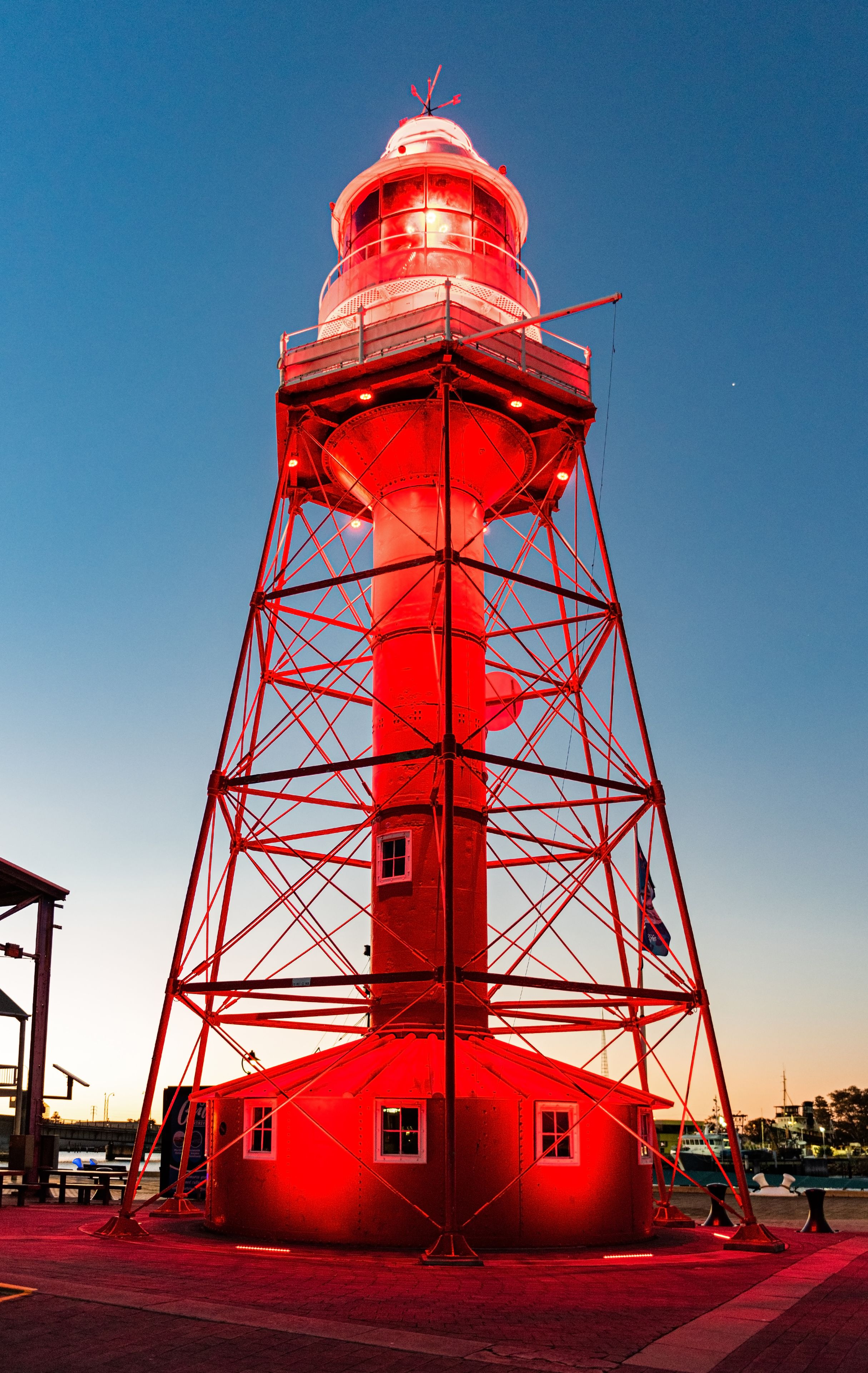 A red lighthouse with a metal framework, a small building at its base, and a clear blue sky in the background.