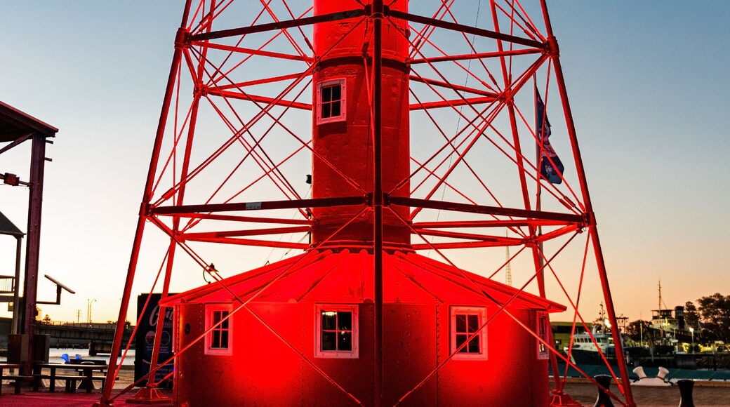 A red lighthouse with a metal framework, a small building at its base, and a clear blue sky in the background.