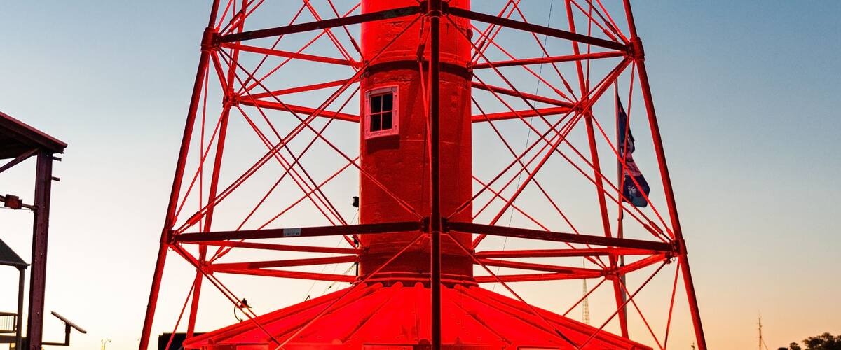 A red lighthouse with a metal framework, a small building at its base, and a clear blue sky in the background.