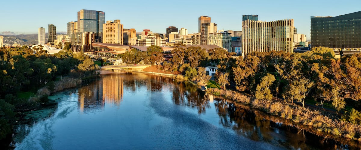 A city skyline with modern buildings, a river reflecting the structures, and a park with trees.
