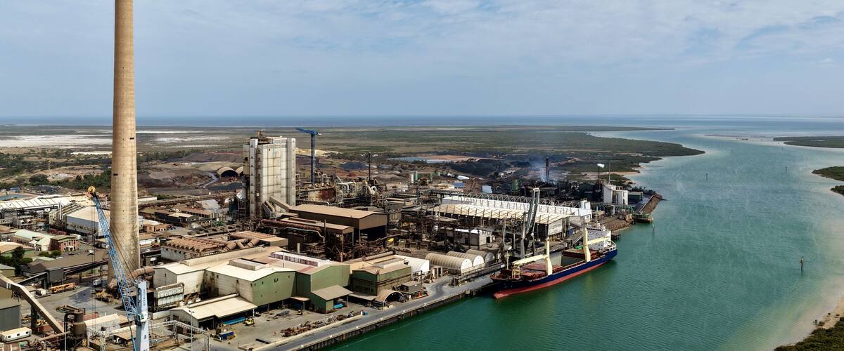 An industrial complex with a tall chimney, a large ship docked at a pier, and a river in the background.
