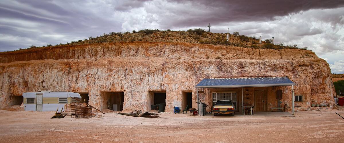 A large rock formation with a building carved into it, a car parked in front, and a trailer in the background.
