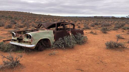 An abandoned, rusted car in a desert landscape with sparse vegetation and a cloudy sky.