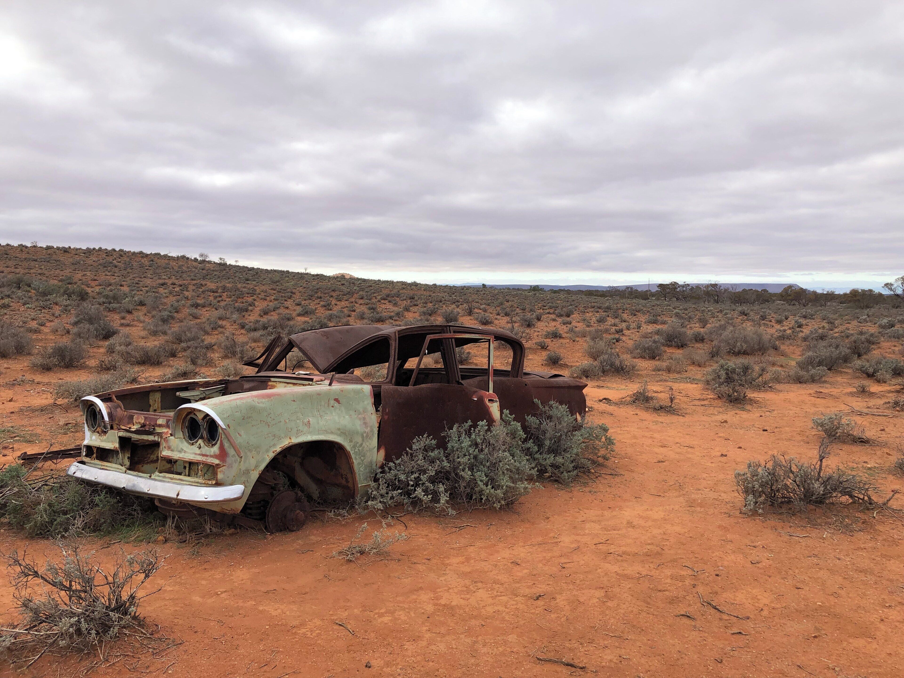 An abandoned, rusted car in a desert landscape with sparse vegetation and a cloudy sky.
