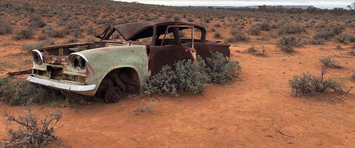 An abandoned, rusted car in a desert landscape with sparse vegetation and a cloudy sky.