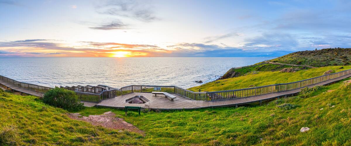 Vista de un atardecer costero con un paseo de madera y terreno cubierto de hierba.