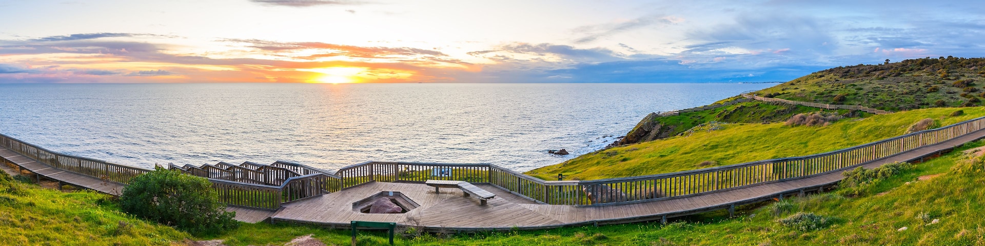 Vista de un atardecer costero con un paseo de madera y terreno cubierto de hierba.