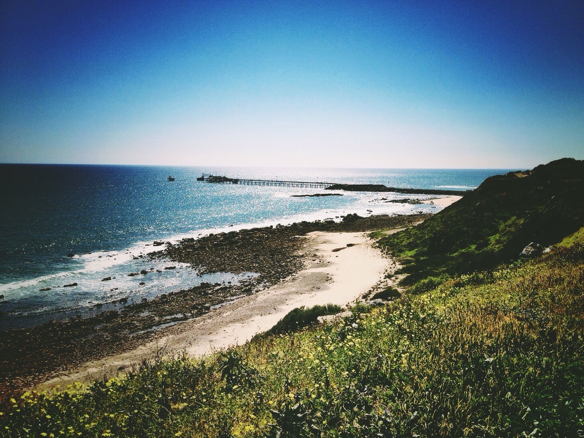 Paisagem costeira com uma praia arenosa, falésias rochosas e um cais que se estende para o oceano.