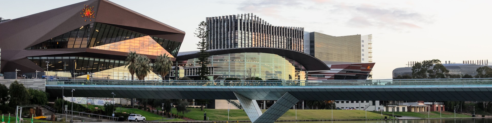 Eine moderne Brücke über ein Gewässer mit Spiegelungen, ein markantes Gebäude mit einem besonderen Dach und ein weiteres Gebäude mit einer strukturierten Fassade.