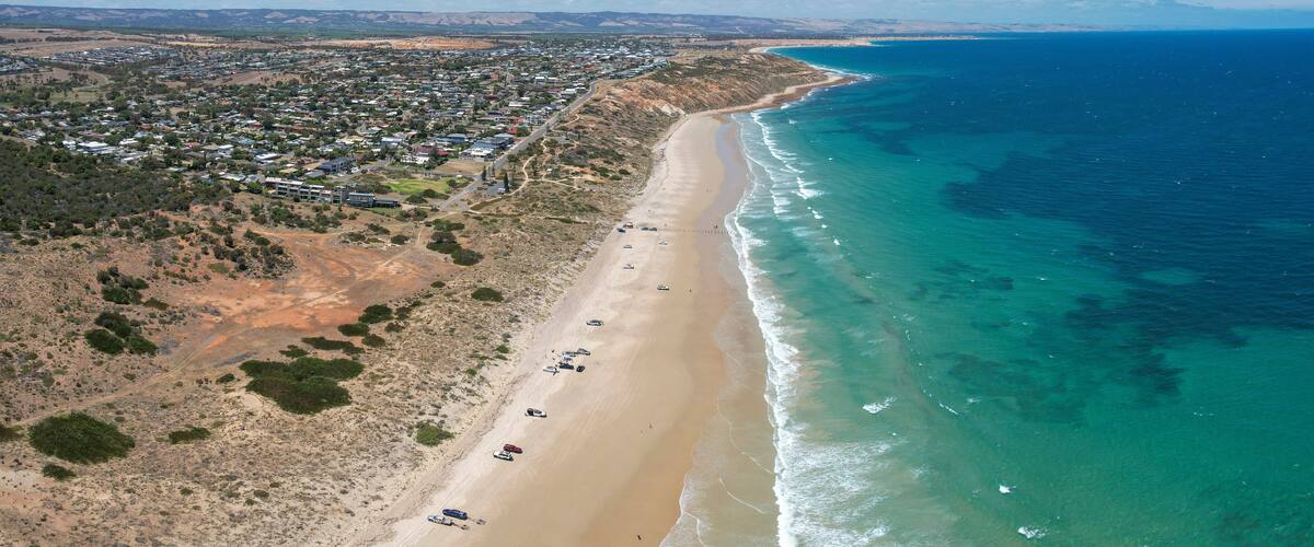 Vista aérea de un pueblo costero con una extensa playa de arena, mar azul cristalino y una zona interior montañosa con edificios dispersos.