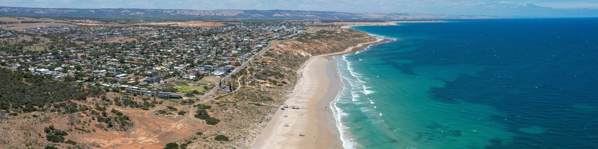 Vista aérea de un pueblo costero con una extensa playa de arena, mar azul cristalino y una zona interior montañosa con edificios dispersos.