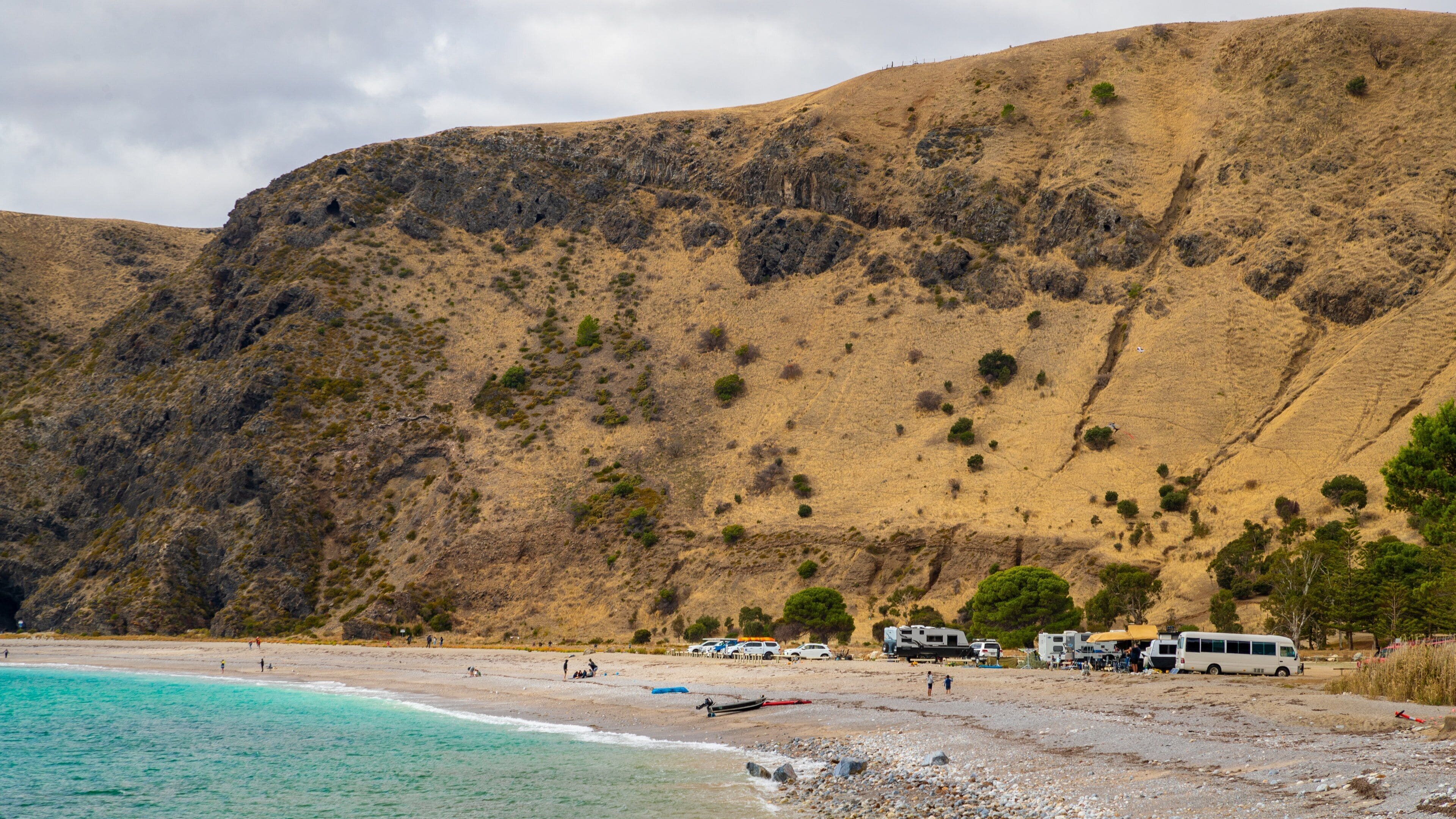A beach with clear turquoise waters, a rocky hillside, and several RVs parked on the sand.
