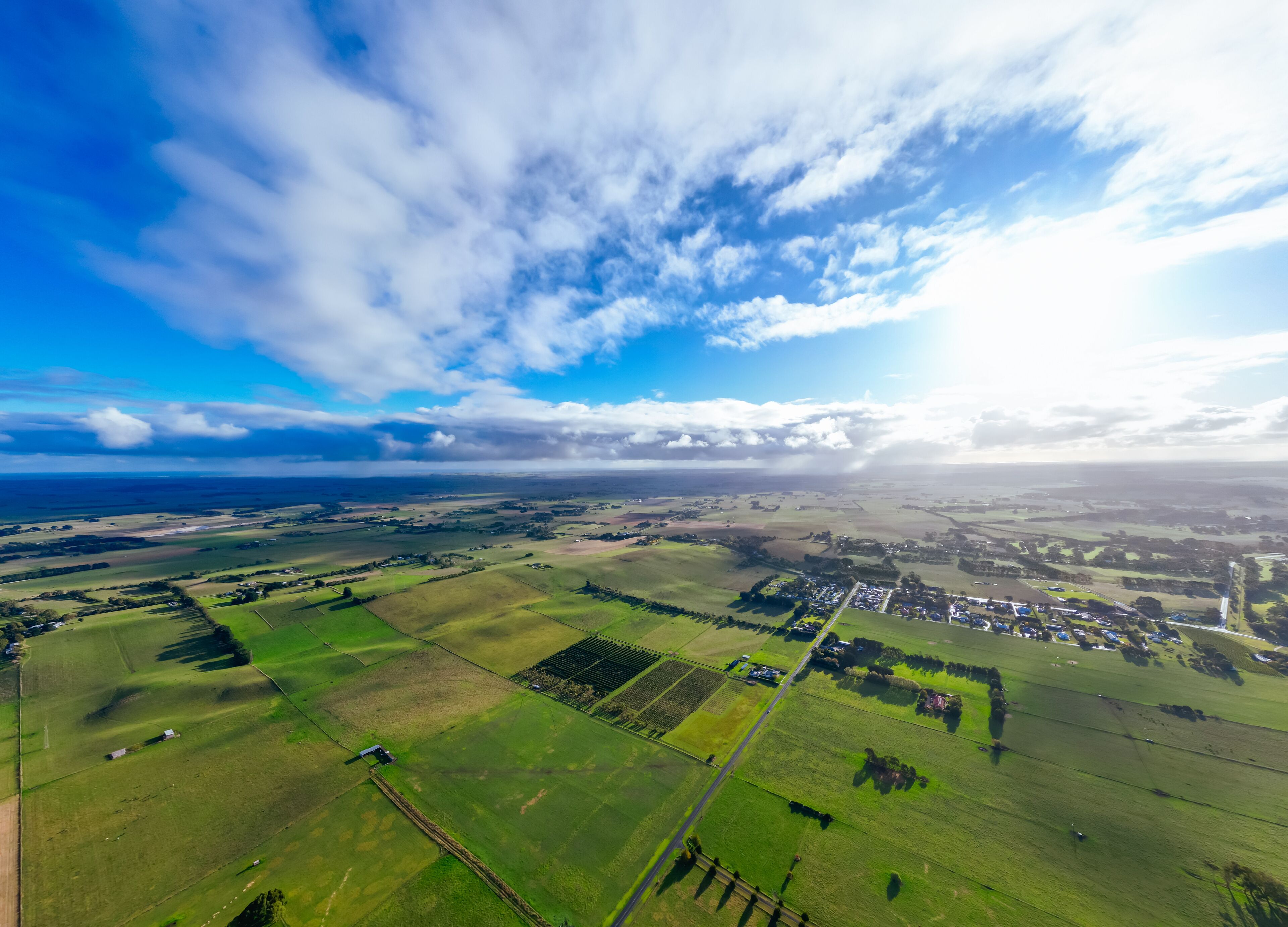 A rural landscape with fields, a small village, and a distant coastline under a partly cloudy sky.