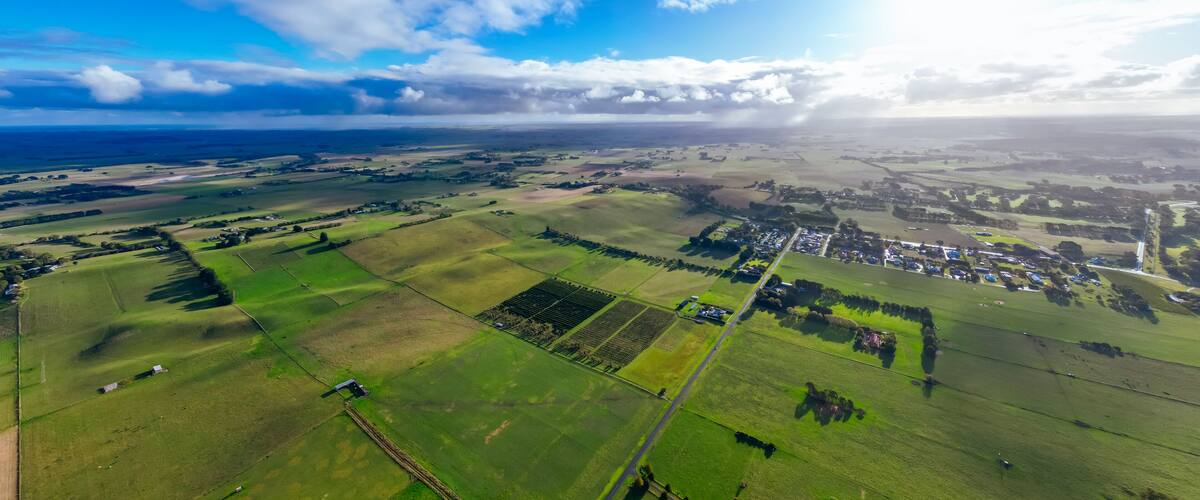 A rural landscape with fields, a small village, and a distant coastline under a partly cloudy sky.
