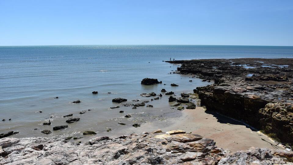 A rocky shoreline with scattered rocks in the water and a clear blue sky.