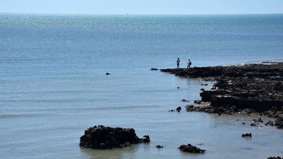 A rocky shoreline with two people fishing, calm sea, and clear sky.